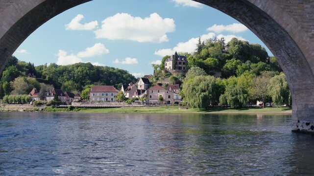 Unique perspective of Limeuil village in Dordogne, France, viewed through a stone bridge arch. The river flows in the foreground with the historic hilltop town in the background.