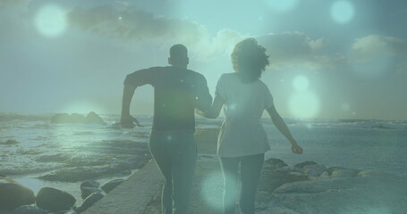 Image of spots of light over african american couple running on the rocks near the sea