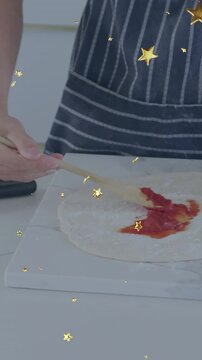 Vertical video: Teen cook bringing spoon to dough smearing sauce for pizza as gold stars over hands