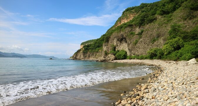 A beach on the island of Sumbawa, Indonesia, which has beautiful views and consists of blue sea with rocky beaches and towering rocky hills with dense green vegetation.
