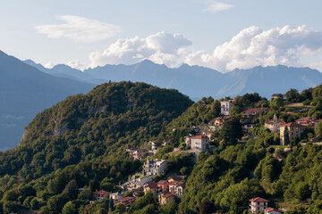 A picturesque hillside town nestled amongst lush green trees with a dramatic backdrop of distant mountains under a cloudy sky