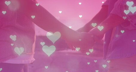 Image of multiple heart icons falling over mid section of couple holding hands near the sea