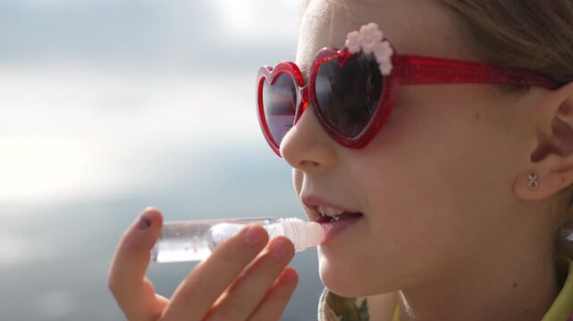 Girl, sunglasses, lip gloss, applying shimmering cosmetic makeup outdoors on a sunny summer day.