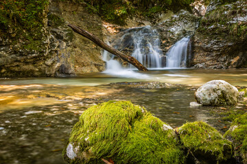 Water flows over rocks and moss in Mostnica Gorge near Stara Fuzina. Green moss covers stones near the water. Trees surround the area, adding to the natural scenery.