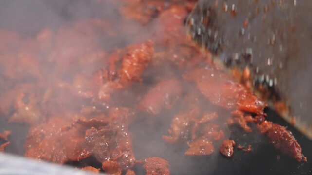 POV close-up of sizzling al pastor meat being cooked with a spatula and knife on a hot Mexican street food grill. Highlights aroma, texture, and savory, flavorful experience.