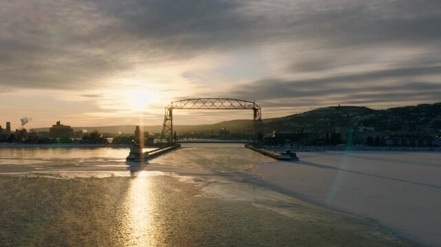 Cinematic drone push through the Duluth Ship Canal toward the iconic Aerial Lift Bridge at sunset. Thick sea smoke and golden light create an ethereal winter maritime scene