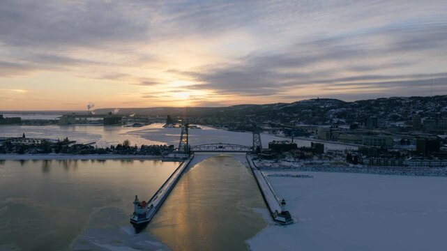 High-altitude panoramic aerial of the Duluth harbor and Lift Bridge during an arctic sunset. Sea smoke swirls over the frozen lake while city lights begin to glow.
