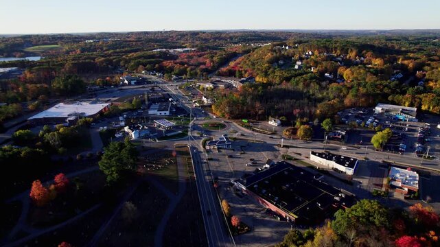 Early morning traffic activity on a traffic rotary with surrounding roadways and commercial sites. Sprawling New England fall foliage and woodland.