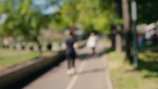 Out of focus shot of a park pathway with people enjoying a sunny day, creating a beautiful bokeh background effect with green trees and sunlight for a calm, abstract and dreamy atmosphere