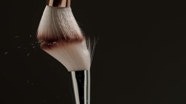 Slow motion clip of two cosmetic brushes colliding in mid-air, creating a cloud of face powder against a dark background, symbolizing makeup application and beauty concepts