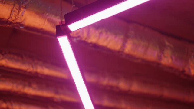 Modern interior design with a hanging geometric fixture of glowing pink led light tubes swinging slowly. Illuminating an industrial-style ceiling with reflective pipes and creating a futuristic vibe