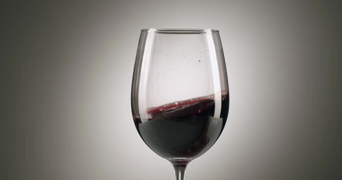 Elegant glass of red wine being swirled in a circular motion against a smooth, graduated gray background, creating beautiful waves and highlighting the deep color and viscosity of the beverage