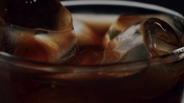 Close-up of black coffee pouring into a glass filled with ice cubes against a dark background, making a refreshing cold beverage perfect for a hot summer day in slow motion