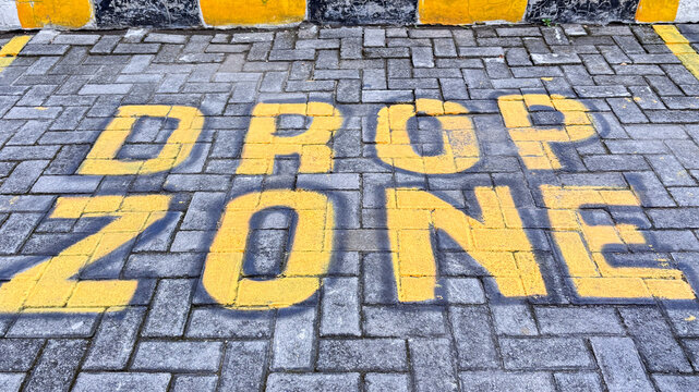 Top view of "DROP ZONE" text painted in yellow stenciled letters on a grey paving block surface in a parking area or roadside.
