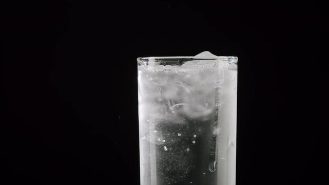 Close-up of an effervescent pill dissolving in a glass of sparkling water with ice cubes against a black background. White swirls of gas and bubbles rise, creating a fizzy, refreshing drink