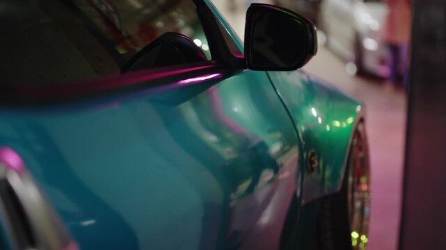 Close-up side view of a modern sports car with a stunning teal and green chameleon paint finish, reflecting vibrant pink and green lights at a custom auto show in a dimly lit environment