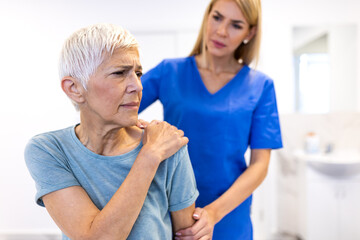 Obraz premium Pain in the shoulder. Upper arm pain, people with body-muscles problem, healthcare and medicine concept. Senior woman sitting in clinic and holding painful shoulder while nurse or doctor helps her.