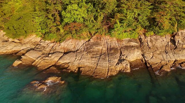 Aerial cinematic drone flyaway from rocky lava coast of Phuket, Thailand. Waves crash against tropical shoreline and flow into clear, transparent waters of the Andaman Sea