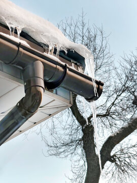 Icicles hanging from house gutter with snow on roof edge against clear winter sky and bare tree branches outdoors.
