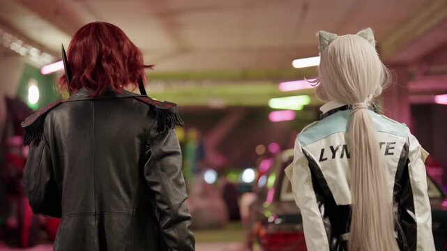 Rear view of two female cosplayers walking through a brightly lit parking garage during a popular culture event, showcasing their elaborate costumes and hairstyles in slow motion
