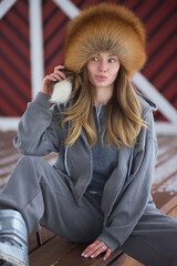 Fototapeta premium Happy woman adjusting her cozy winter hat. Woman in relaxed attire smiles in rustic cabin setting. Natural shot of cheerful woman fixing her warm fur hat outdoors