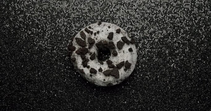 Delicious white frosted donut with chocolate cookie crumble topping slowly spinning on a textured black background, showcasing a popular sweet treat from a top-down perspective