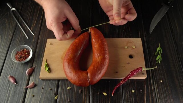 Hands of a person tying a sausage with green threads on a wooden cutting board surrounded by spices and garlic cloves in a kitchen setting