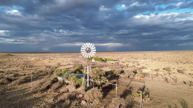 The camera rises up in front of a fast-spinning wind pump on a semi-arid landscape, with stormy clouds. 4K Aerial Video.