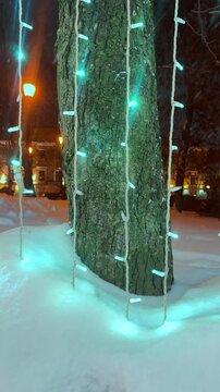 The vertical video shows a city street at night, where garlands on trees, lampposts, and buildings shimmer with multicolored lights through the fresh snow. 