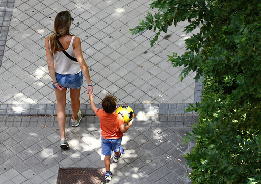 Madre con hijo de la mano con bal&oacute;n paseando en verano camino del parque a jugar