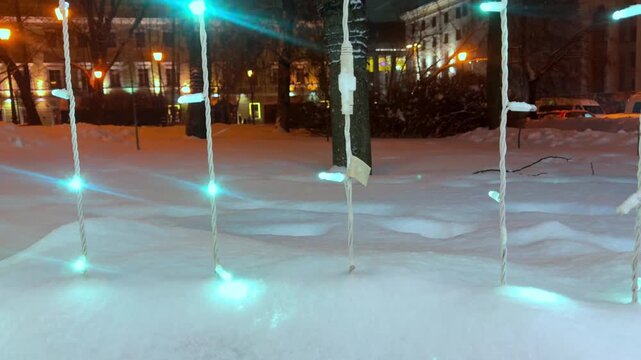 The vertical video shows a city street at night, where garlands on trees, lampposts, and buildings shimmer with multicolored lights through the fresh snow. 
