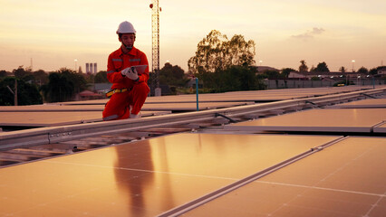 An Asian solar technician in an orange jumpsuit points toward the horizon on a rooftop at sunset. Clean energy vision, sustainable development, and professional solar installation