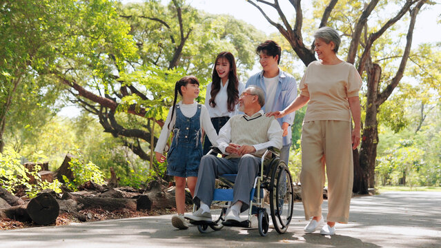 An Asian family enjoys a walk in a lush green park, supporting a senior man in a wheelchair. Multigenerational family authentic love, care, accessibility, and happiness outdoors