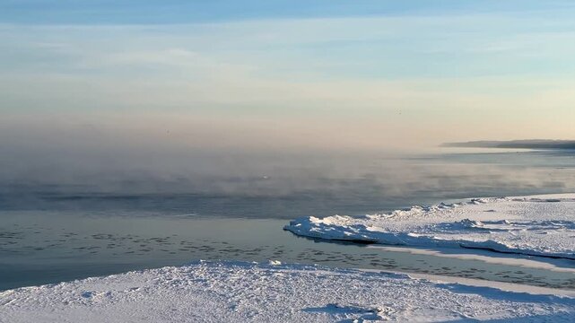 A winter morning seascape unfolds a frozen shoreline, where layers of snow and ice stretch out toward a misty water with cold fog mooving