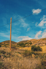 Obraz premium Wooden electricity post in arid rocky hilly landscape with some vegetation and trees under a blue sky with some clouds. Huebro, Almeria, Andalusia, Spain.