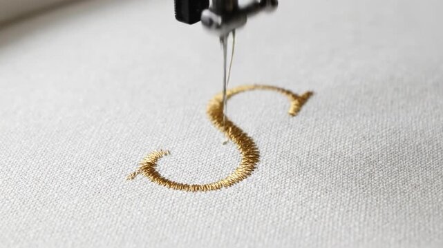 Watch the mesmerizing process as an embroidery machine uses bright gold thread to precisely stitch the capital letter 'S' onto a crisp white textile surface, showcasing skilled craftsmanship.