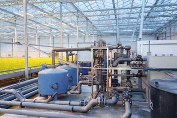 Fototapeta na wymiar An extensive system of industrial pipes, pumps, and water treatment tanks supports hydroponic plant growth within a vast, glass-roofed commercial greenhouse during daylight hours.