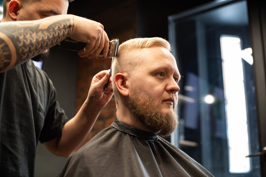 Barber cutting a man&rsquo;s hair with clippers in a barbershop. Side profile of a male client with short haircut and beard, professional grooming service in a salon.