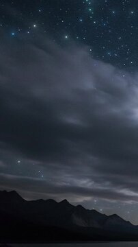 Gazing at stars and slow-moving clouds over dark mountain silhouettes at night, mirrored in a tranquil lake in the wilderness. Looping animation.