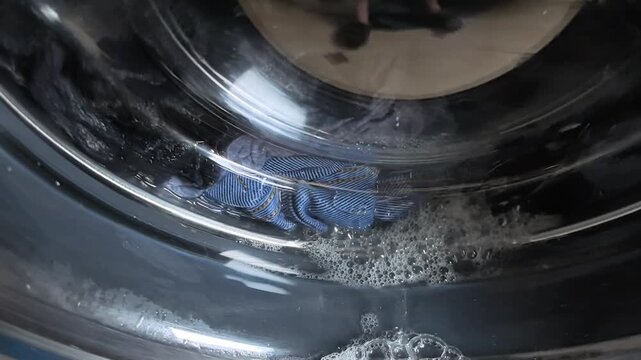 The washing machine is cleaning clothes with soap and water in a home laundry room. Bubbles form in the glass door as the cycle runs. The setting shows a typical weekday afternoon.