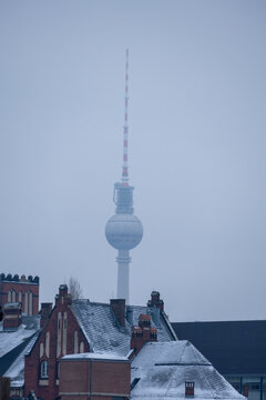 Berlin TV Tower rises above snowy rooftops in winter haze, its sphere and antenna fading into the cold gray sky over the quiet cityscape