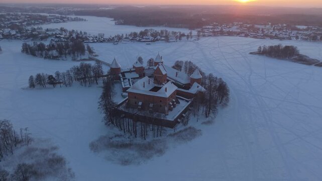 4K aerial drone pull-up over Trakai Island Castle, Lithuania, snow-covered medieval fortress on frozen Lake Galvė with warm winter sunset on horizon, cinematic establishing shot