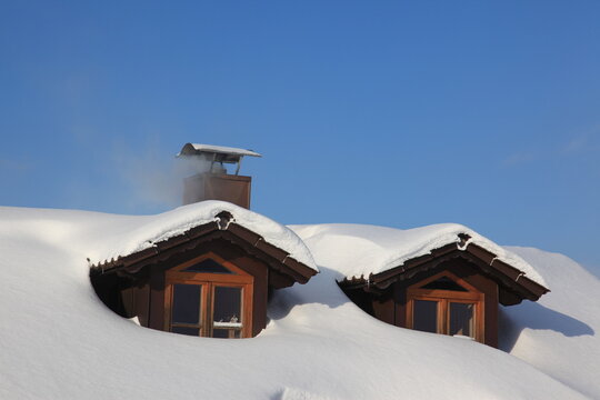 Snow-covered dormer windows with thick white layers on the roof, smoke gently rising from the chimney against a clear winter sky.
