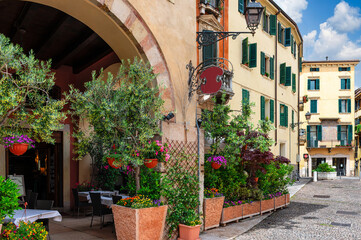 Fototapeta premium Old street with flowers and tables of restaurant in Verona, Italy. Architecture and landmark of Verona. Cozy cityscape of Verona.