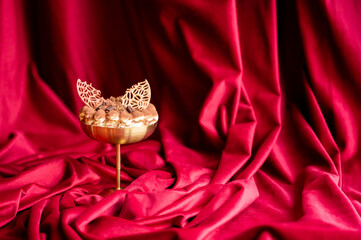 Obraz premium Elegant gourmet tiramisu dessert served in a gold coupe glass, topped with chocolate coffee bean garnishes and cocoa powder, set against a luxurious, draped red velvet fabric background