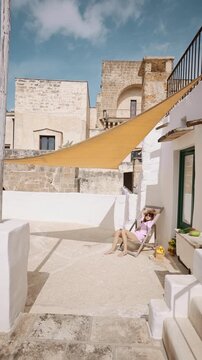 Static video of a woman relaxing on a sunlit terrace in southern Italy. A light breeze gently moves the sun shade, creating a calm, peaceful atmosphere of Mediterranean slow living and everyday summer