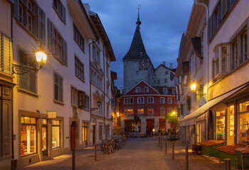 Obraz premium Early morning scene in Zurich old town with warm lights, empty street, traditional buildings, and a historic clock tower at sunrise