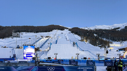 Obraz premium Livigno, Italy - February 6, 2026: Freestyle skiing venue with massive snow jump and competition course prepared for Winter Olympic Games in Livigno under clear blue sky
