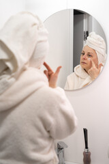 Fototapeta premium Woman in bathrobe and towel applying moisturizing eye cream during daily beauty routine in bathroom