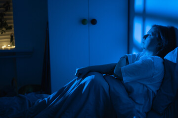 Fototapeta premium Exhausted middle-aged woman sitting in bed and looking through the window at night, female suffering from insomnia and work stress feeling lonely and sleepless